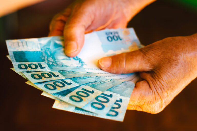 As mãos de uma pessoa segurando cinco cédulas de 100 reais, espalhadas em um fundo desfocado, possivelmente representando o benefício fgts: trabalhador poderá efetuar saque emergencial.