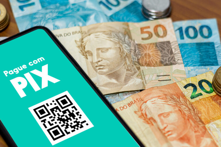 A tela de um smartphone exibe o Pague com PIX com um código QR. Cédulas de real - 50, 100, 20 - e moedas estão espalhadas em uma mesa ao fundo. Atenção: você tem 90 dias como limite para receber o prêmio.