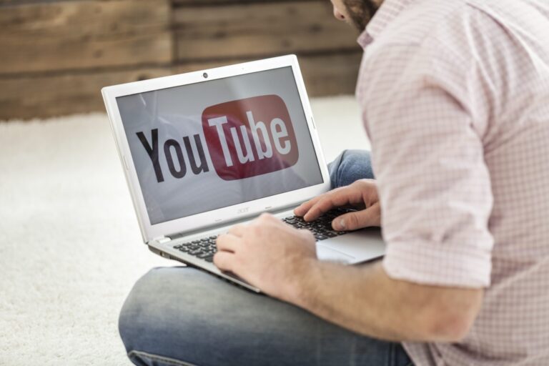 Uma pessoa com uma camisa xadrez rosa senta-se de pernas cruzadas no chão, usando um laptop com o logotipo do canal YouTube lotéricos exibido na tela.