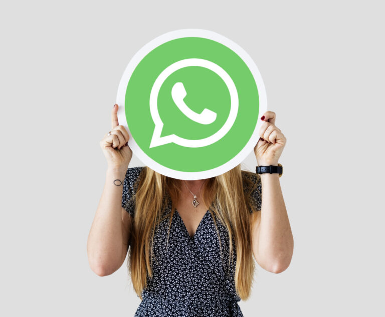 Uma mulher com longos cabelos loiros segura um grande cartaz redondo com o logotipo verde do WhatsApp em frente ao seu rosto, em um fundo cinza claro, como se estivesse ponderando se vale a pena usar o WhatsApp.