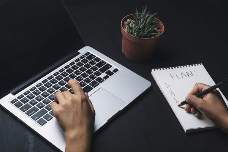 Uma pessoa digita em um laptop com uma das mãos enquanto escreve uma lista numerada intitulada PLAN em um bloco de notas com a outra - talvez um brainstorming de copywriting para venda de jogos em lotérica. Perto dali, um pequeno vaso de suculentas repousa sobre a mesa escura.