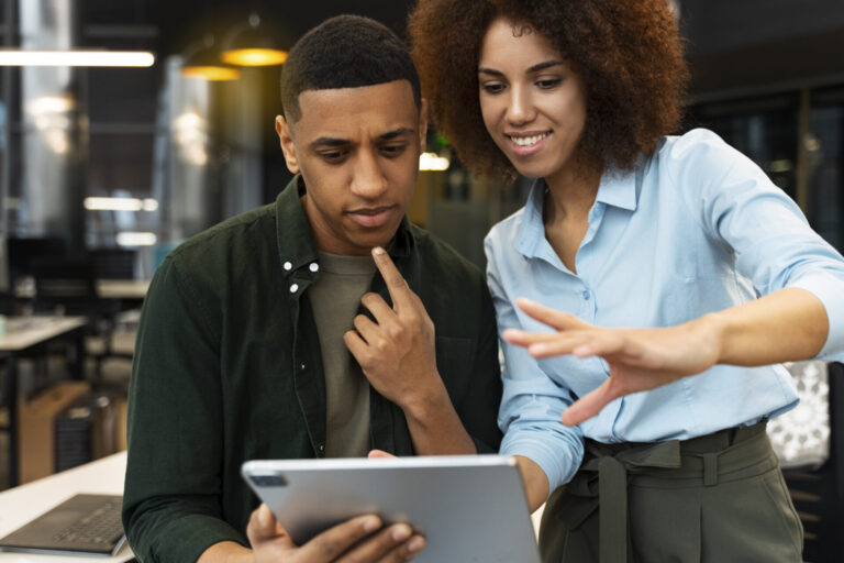 Dois jovens adultos, um homem e uma mulher, olham juntos para um tablet em um escritório moderno. O homem parece pensativo, tocando o queixo, enquanto a mulher sorri e faz gestos, explicando como iniciar um projeto ou fazer consultoria.
