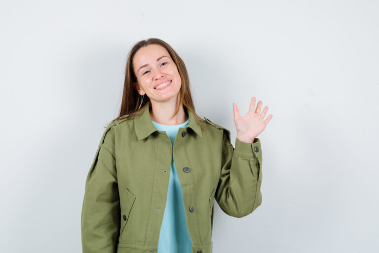 Uma jovem de longos cabelos castanhos, vestindo uma jaqueta verde e camisa azul-clara, sorri e acena com a mão direita como se estivesse cumprimentando alguém em uma casa lotérica, contra um fundo liso e de cor clara.
