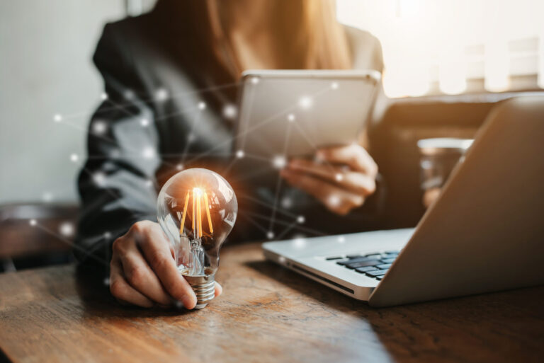 Uma pessoa de terno segura uma lâmpada brilhante em uma mão e um tablet na outra, com um laptop na mesa, simbolizando inovação, tecnologia e assim análise 360° no ambiente de trabalho.