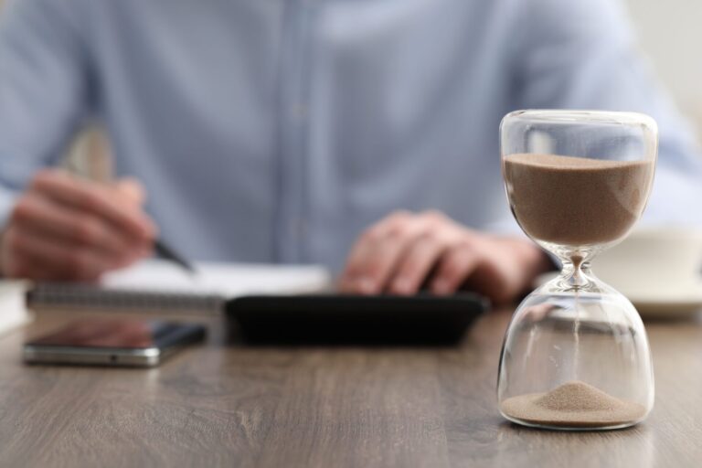Uma ampulheta, que simboliza a gestão de tempo, fica em uma mesa de madeira em primeiro plano, enquanto uma pessoa escreve em um caderno, com um smartphone e uma caneta por perto - ferramentas para organizar melhor e lucrar com eficiência.