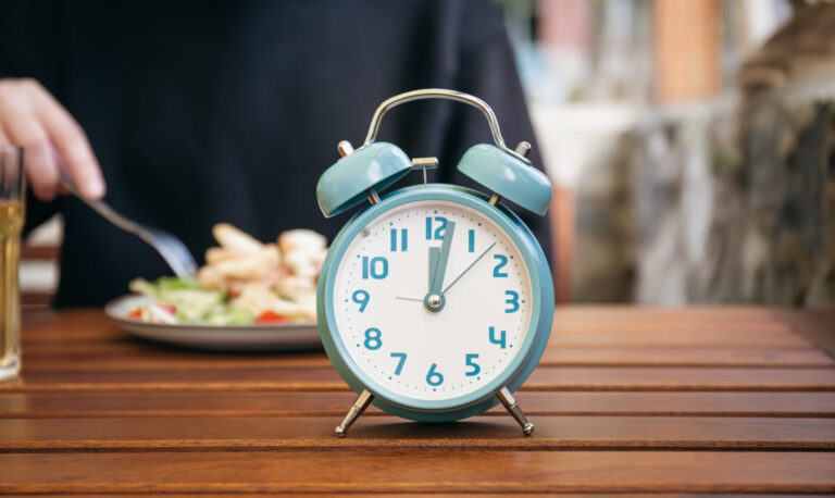 Um despertador azul mostrando 12:00 está sobre uma mesa de madeira. Ao fundo, enquanto alguém faz uma refeição, a cena sugere passos para organizar o horário, com um prato de comida e um garfo fora de foco.