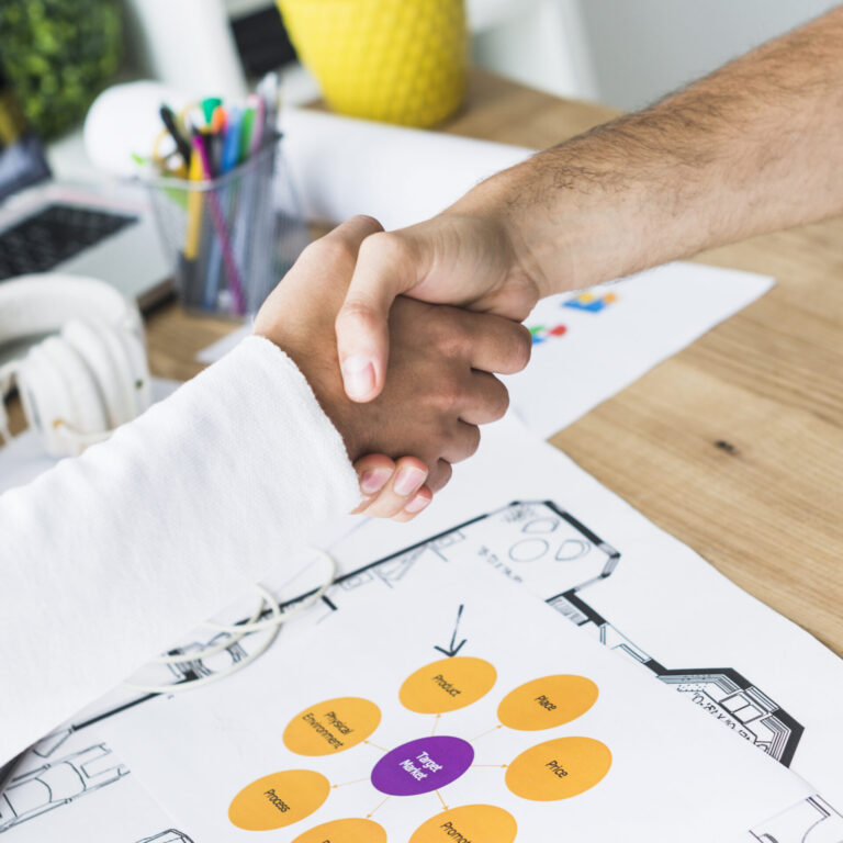 Duas pessoas se cumprimentam sobre uma mesa com documentos, gráficos e materiais de escritório, sugerindo um acordo bem-sucedido que atende às necessidades de uma obra ou parceria comercial.
