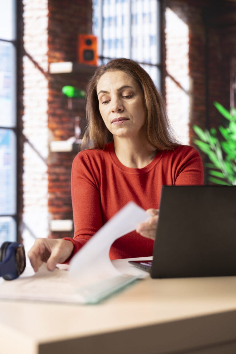 Uma mulher com um suéter vermelho senta-se em uma mesa com um laptop, olhando para papéis em sua mão. Ela parece concentrada, possivelmente encerrando o expediente mais cedo, em um escritório moderno com grandes janelas e prateleiras ao fundo.