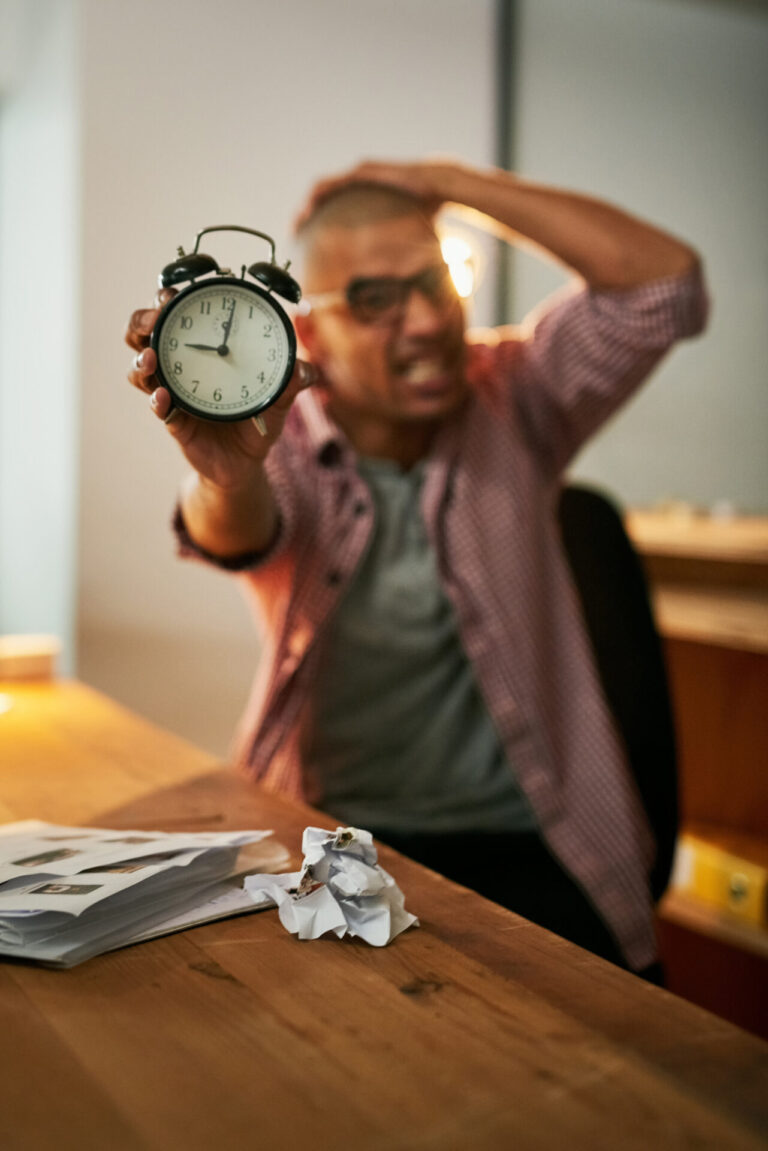 Uma pessoa estressada está sentada em uma mesa, segurando um despertador em direção à câmera, como se estivesse perguntando se existe limite de horas. A outra mão está sobre a cabeça, cercada de papéis e folhas amassadas sobre a mesa de madeira.