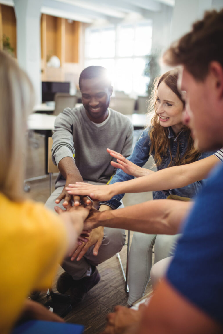 Um grupo de pessoas sentadas em círculo, sorrindo e colocando as mãos no centro, demonstrando trabalho em equipe e união em um ambiente de escritório moderno e bem iluminado, com foco em ações sociais que beneficiam a comunidade.