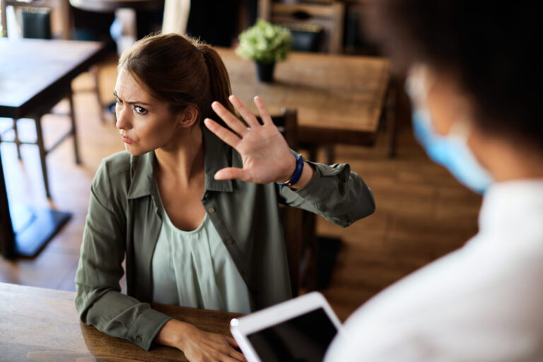 Uma mulher sentada à mesa de um restaurante levanta a mão, sinalizando "pare" para alguém com um tablet. Com expressão de irritação, ela mostra claramente ser uma cliente nervoso-saiba como lidar com situações delicadas assim.