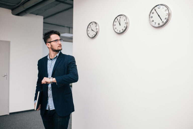 Um homem de blazer e óculos verifica seu relógio de pulso enquanto segura uma pasta, ao lado de uma parede branca onde três relógios exibem horários diferentes - um cenário ideal para criar quadros horários para programações globais.