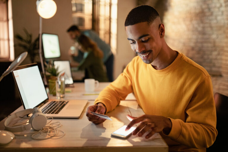 Um homem sorridente com um suéter amarelo está sentado em uma mesa usando seu smartphone e segurando um cartão de crédito, talvez explorando 3 aplicativos inovadores que podem mudar sua rotina. Um laptop, fones de ouvido e uma xícara estão próximos no fundo do escritório moderno.