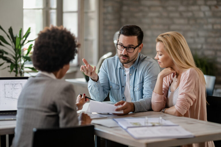 Um homem e uma mulher sentam-se em uma mesa em frente a uma mulher profissional, discutindo documentos. O homem faz gestos enquanto fala, enfatizando que a dicção fundamental é crucial nesta hora. Papéis e um laptop com uma planta baixa estão sobre a mesa nesse ambiente de escritório.