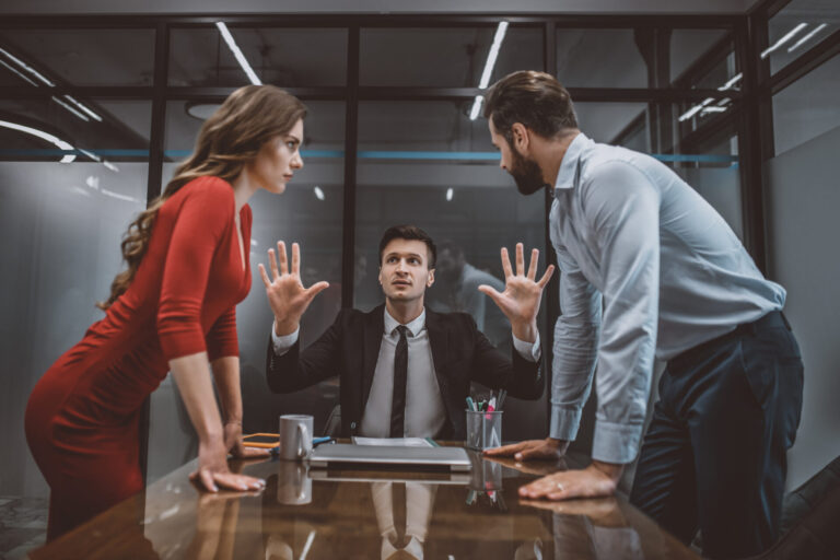 Um homem de terno senta-se em uma mesa com as mãos levantadas, parecendo mediar conflitos no ambiente de trabalho: como lidar entre uma mulher de vestido vermelho e um homem de camisa azul, ambos tensos e inclinados para frente em um escritório moderno.