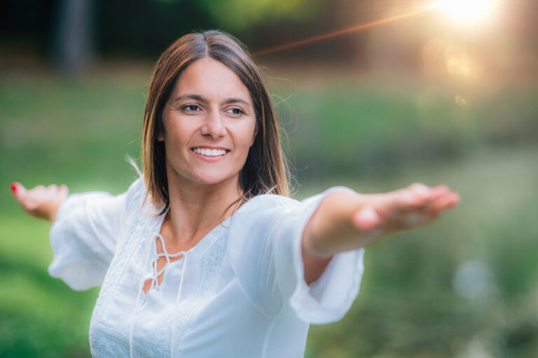 Uma mulher vestindo uma blusa branca pratica ioga ao ar livre com os braços estendidos, sorrindo e olhando para o lado. A luz do sol brilha ao fundo, lembrando-nos de que o equilíbrio emocional é fundamental para casas lotéricas.