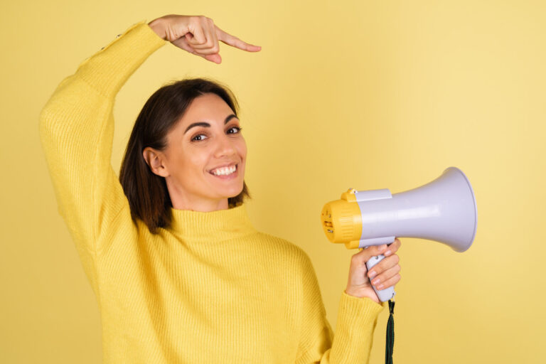 Uma mulher sorridente com um suéter amarelo segura um megafone e aponta para ele, em pé diante de um fundo amarelo - demonstrando o apelo à ação na prática: como levar sua mensagem de forma impactante.
