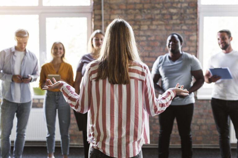 Uma pessoa com cabelos longos e camisa listrada está de braços abertos, de frente para um grupo de cinco pessoas sorridentes em um ambiente fechado, com a luz do sol entrando pelas grandes janelas - destacando a importância de registrar talentos em um ambiente acolhedor.
