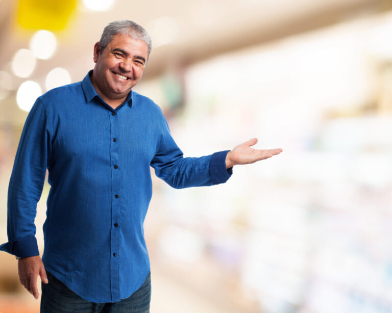 Um homem sorridente de camisa azul está em um ambiente interno, gesticulando com a mão direita como se estivesse apresentando 6 livros que todo bom vendedor precisa. O fundo está bem iluminado e desfocado.