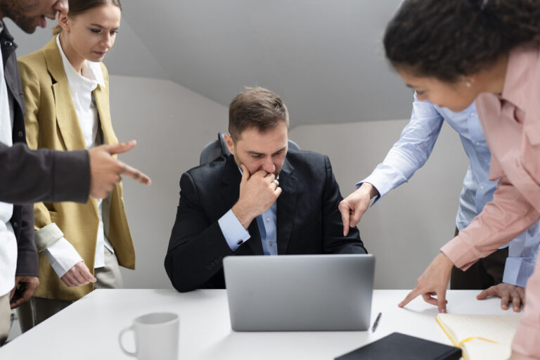 Um homem está sentado em uma mesa, parecendo preocupado enquanto trabalha em um laptop, enquanto três colegas estão ao seu redor apontando para a tela, sugerindo uma situação de trabalho tensa e a necessidade de estabelecer limites proprietários.
