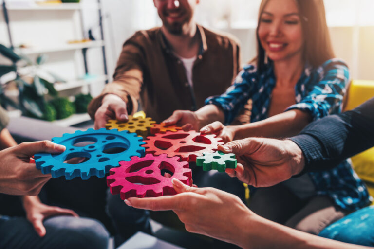 Um grupo de pessoas sorrindo e segurando engrenagens coloridas entrelaçadas, simbolizando o trabalho em equipe e a colaboração em um ambiente de escritório bem iluminado e casual que ajuda a definir a cultura organizacional.