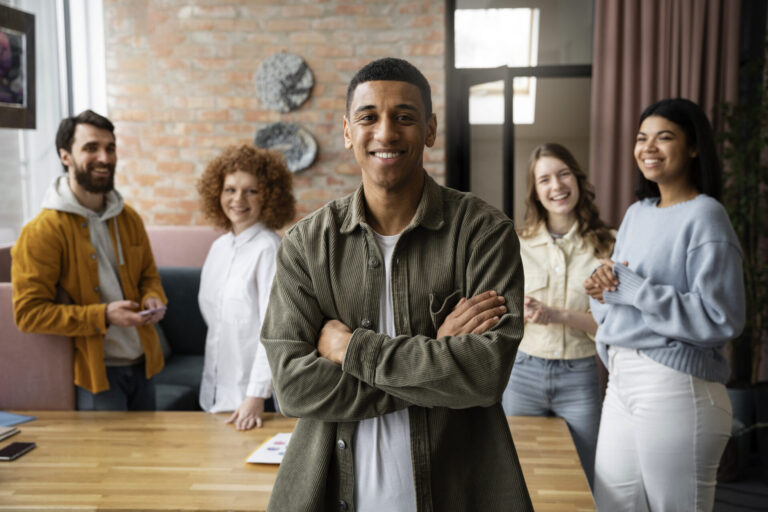 Um jovem está confiante com os braços cruzados em primeiro plano, sorrindo para a câmera. Quatro colegas da equipe do departamento pessoal casas estão atrás dele, perto de uma mesa, também sorrindo, em um ambiente de escritório moderno e bem iluminado.