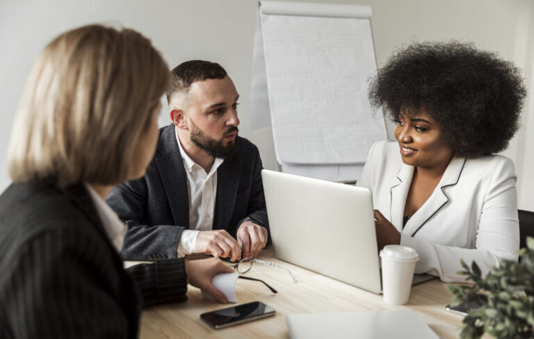Três pessoas em trajes de negócios sentam-se ao redor de uma mesa com um laptop, papéis e café, envolvidos em consultoria para uma equipe lotérica. Um flip chart em branco é visível ao fundo enquanto eles discutem estratégias.