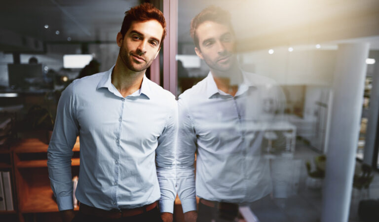 Um homem vestindo uma camisa azul clara fica em um ambiente interno, encostado em uma parede de vidro com seu reflexo visível - talvez ponderando se é um gestor ou líder qual é o melhor, enquanto sorri levemente no ambiente moderno do escritório.