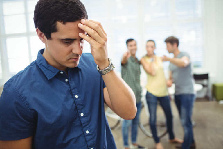 Um homem de camisa azul parece chateado e segura a testa, provavelmente enfrentando um funcionário problema: como lidar, enquanto três pessoas ao fundo apontam e riem dele em um ambiente interno iluminado.