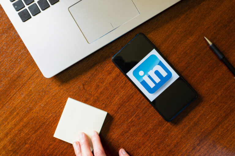 Um smartphone exibindo o logotipo do LinkedIn fica em uma mesa de madeira ao lado de um teclado de laptop, um bloco de notas e uma caneta - perfeito para explorar se LinkedIn para casas lotéricas: vale a pena investir na presença digital do seu negócio.