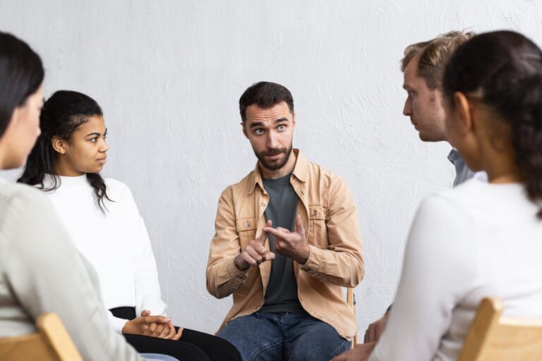Um homem está sentado em um grupo de cinco pessoas, falando com retórica e gesticulando com as mãos enquanto os outros ouvem atentamente. Eles estão sentados em um círculo em cadeiras de madeira contra uma parede branca lisa.