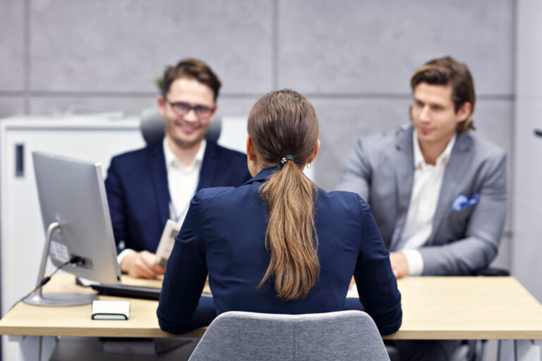 Uma mulher com rabo de cavalo senta-se em frente a uma mesa de dois homens de terno durante uma entrevista de emprego em um escritório moderno, pronta para discutir suas referências profissionais na hora da entrevista, com um computador e documentos sobre a mesa.