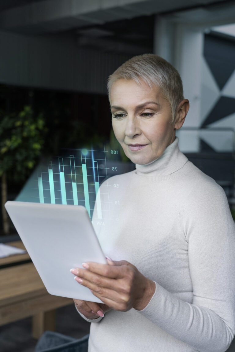 Uma mulher de cabelos grisalhos curtos em uma gola alta clara segura um tablet digital, visualizando um gráfico de barras holográfico. Ela está em um escritório moderno com plantas e decoração geométrica na parede, mostrando como as tecnologias facilitam o ponto no ambiente de trabalho.