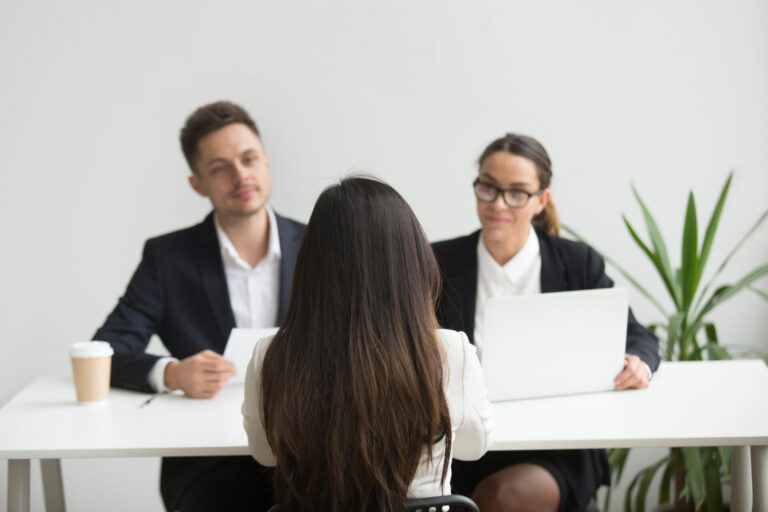 Uma mulher está sentada em frente a dois entrevistadores em trajes de negócios em uma mesa com laptops e uma xícara de café, demonstrando o que levar em conta na hora de contratar em um escritório moderno e iluminado com uma planta ao fundo.