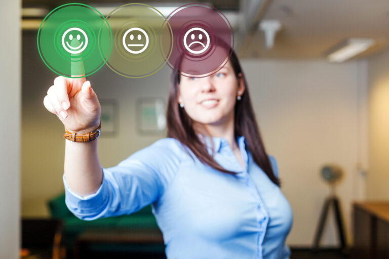 Uma mulher de camisa azul sorri e toca em uma tela virtual, selecionando um emoji de rosto feliz verde entre três rostos, enquanto realiza uma pesquisa de satisfação dos clientes em um ambiente de escritório moderno.