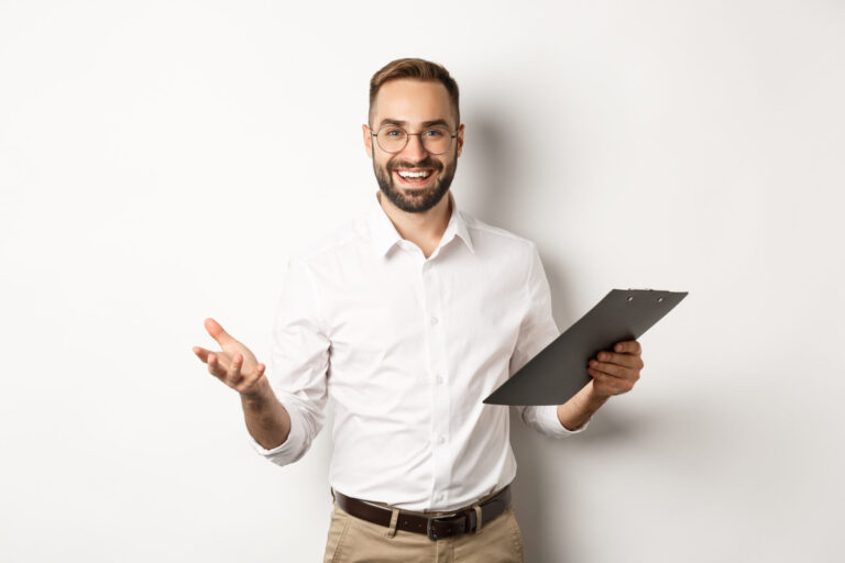 Um homem com barba e óculos, vestindo camisa branca e calça cáqui, está em pé em um fundo branco, sorrindo e segurando uma prancheta, como se estivesse pronto para gerenciar estoques em casa, enquanto gesticula com confiança com a outra mão.