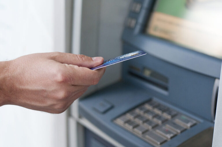 A mão de uma pessoa segura um cartão bancário em frente a um caixa eletrônico, preparando-se para inseri-lo no slot de cartão acima do teclado e da tela, enquanto o caixa comemora anos.