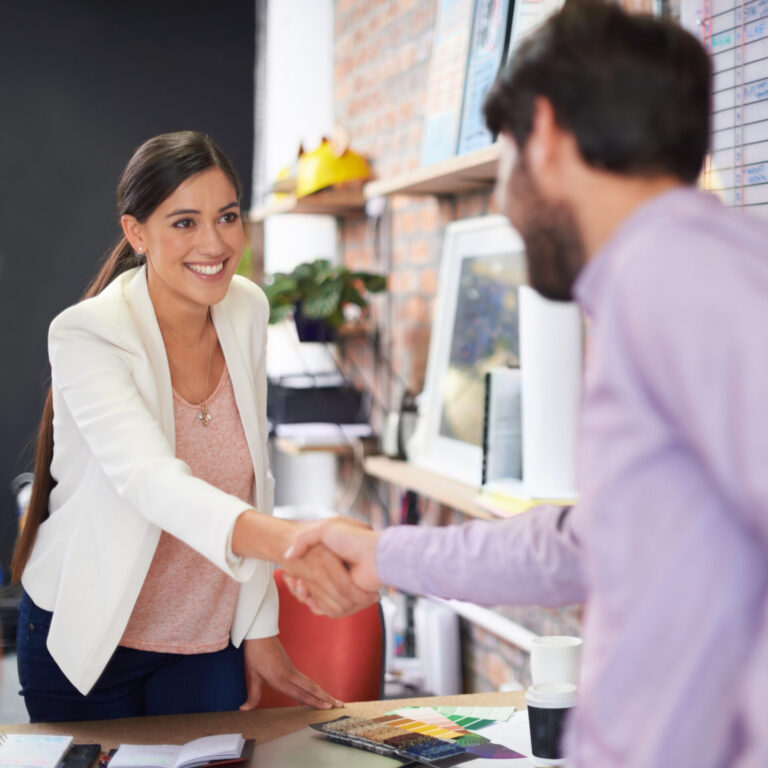 Uma mulher de blazer branco sorri enquanto cumprimenta um homem de camisa roxa clara em uma mesa, possivelmente discutindo como vender meus clientes. A moderna mesa de escritório exibe papéis, um caderno e uma paleta de cores.