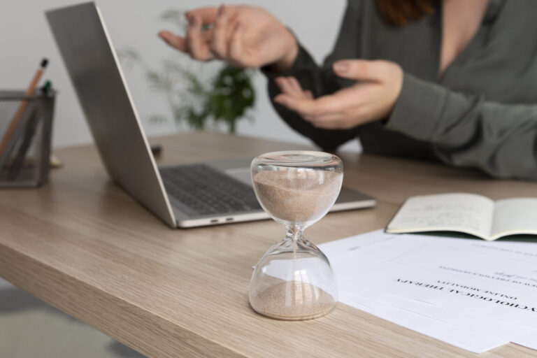 Uma ampulheta em uma mesa em frente a um laptop, um caderno aberto e papéis destaca os gestos de uma pessoa enquanto ela discute uma estratégia de vendas prazo, enfatizando a importância do gerenciamento de tempo nas tarefas diárias.