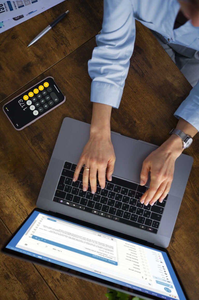Uma pessoa digita em um laptop em uma mesa de madeira, com foco em gerenciamento de contas importantes. Perto dali, um smartphone exibe um aplicativo de calculadora com documentos espalhados. Outra tela de computador está parcialmente visível em primeiro plano.