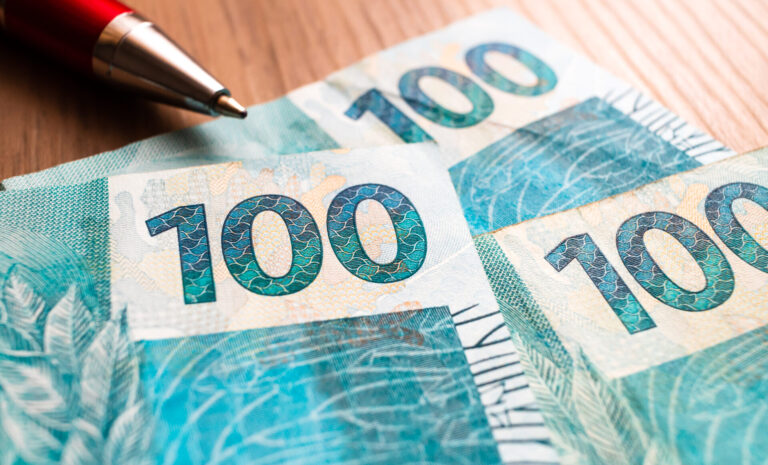Close-up de três cédulas de 100 reais sobre uma superfície de madeira, com parte de uma caneta vermelha visível no canto superior esquerdo - frequentemente vista ao contar o faturamento de lotérica.