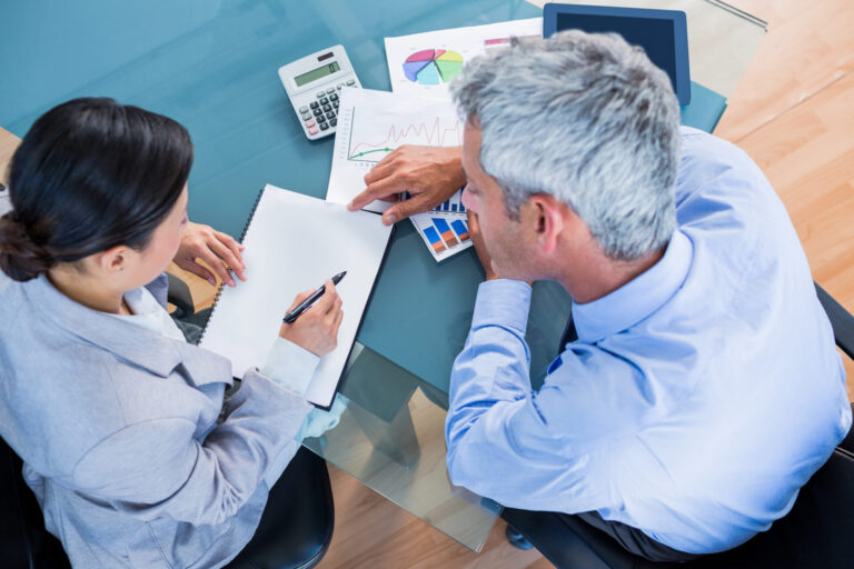 Dois profissionais de negócios sentam-se em uma mesa de vidro com gráficos, uma calculadora e um tablet. Um aponta para um documento enquanto o outro faz anotações, sugerindo uma reunião sobre gestão ou cobrança empresarial.