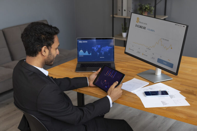 Um homem de terno está sentado em uma mesa de madeira analisando gráficos financeiros em um tablet, com o objetivo de fazer a automação financeira e agilizar processos. Um laptop e um monitor exibem dados de criptomoedas e ações, enquanto papéis e um smartphone também estão sobre a mesa.