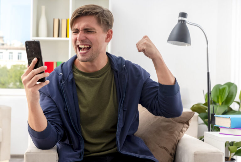 Um jovem sentado em um sofá sorri animado e levanta o punho em comemoração enquanto olha para seu smartphone, refletindo a emoção que alguns sentem com as loterias on-line que põem em risco as casas. Ele usa uma jaqueta escura e camisa verde, com uma luminária, livros e plantas atrás dele.