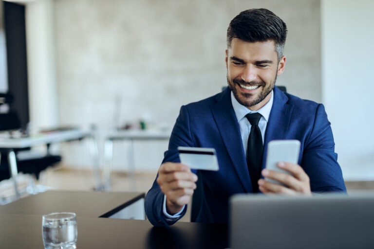 Um homem de terno azul sorri enquanto segura um cartão de crédito e um smartphone em sua mesa, com um laptop e um copo de água à sua frente, gerenciando a cobrança recorrente de cartões em um ambiente de escritório moderno.