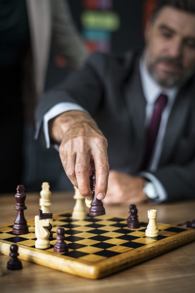 Um homem de terno, incorporando como se estivesse aplicando as 12 regras de ouro para uma liderança, concentra-se em um jogo de xadrez, estendendo a mão para mover uma peça de xadrez preta em um tabuleiro de madeira, enquanto o fundo permanece desfocado.
