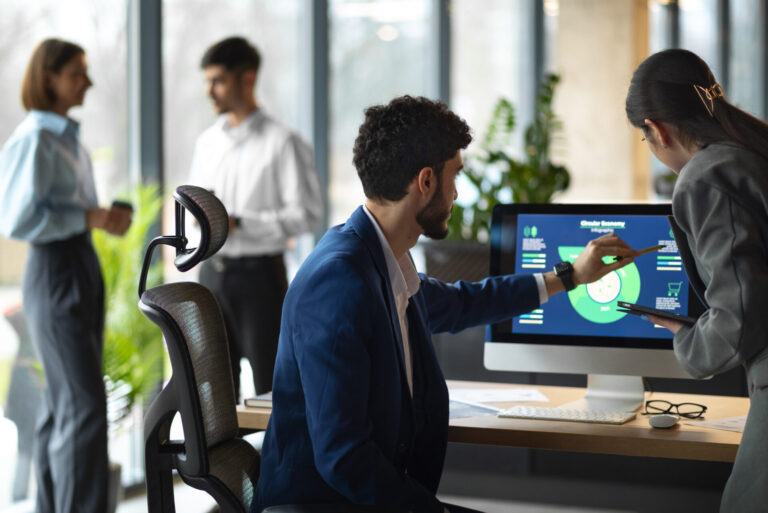 Um homem de terno azul aponta para gráficos de dados que mostram as principais métricas em uma tela de computador enquanto discute com um colega em um escritório. Duas pessoas estão conversando ao fundo, perto de grandes janelas.