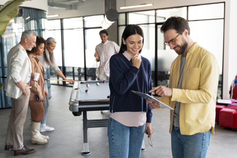 Um grupo de colegas desfruta de um intervalo em um escritório moderno; duas pessoas em primeiro plano discutem a elaboração de um plano de negócio de sucesso em um tablet, enquanto outras jogam bilhar ao fundo.