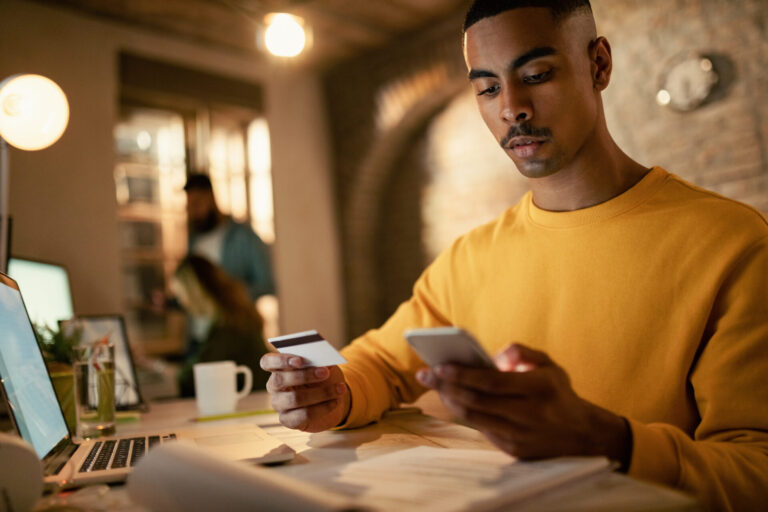Um homem de suéter amarelo está sentado em uma mesa, segurando um cartão de crédito e um smartphone - talvez explorando as melhores plataformas de pagamento. Papéis, um laptop e uma caneca de café repousam sobre a mesa; duas pessoas estão desfocadas no fundo aconchegante e pouco iluminado.