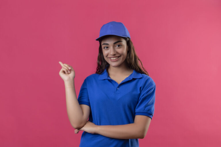 Uma mulher vestindo uma camisa polo azul e boné combinando sorri enquanto aponta para o lado, representando como fazer a padronização de uniformes para sua equipe, em pé contra um fundo rosa sólido.